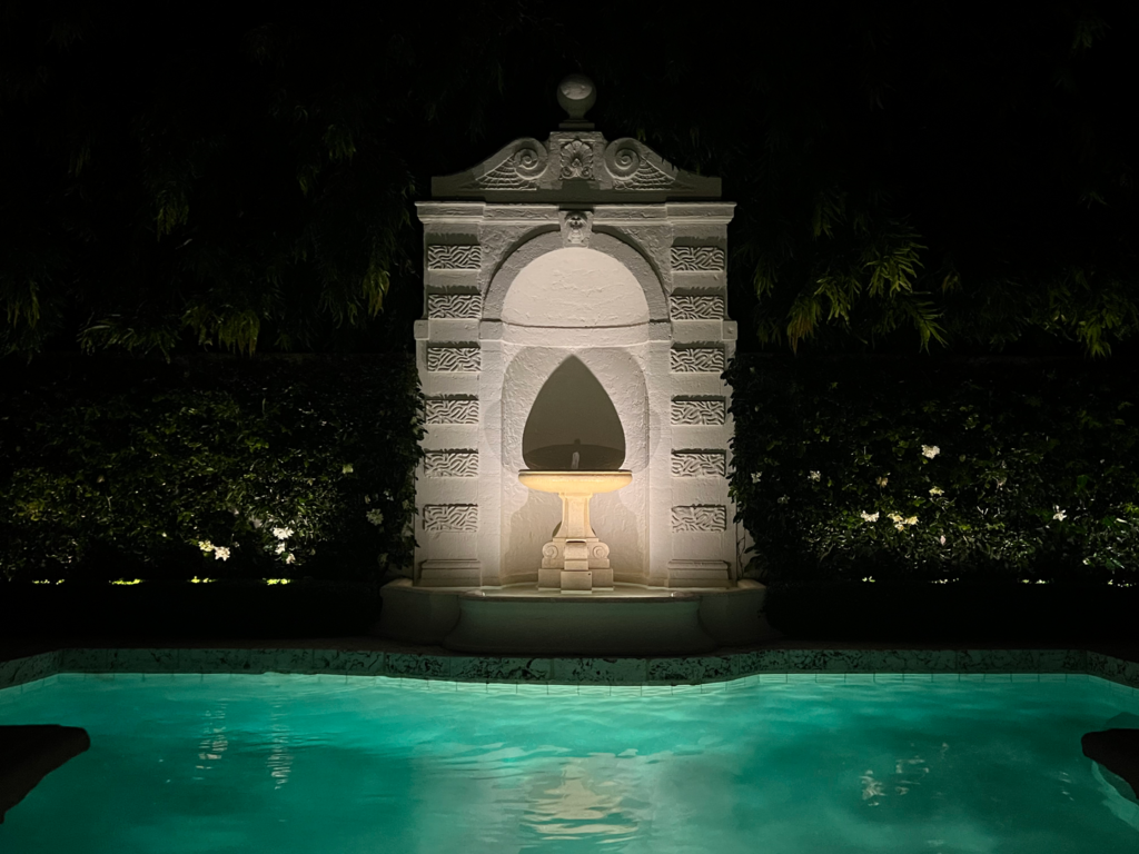 Nighttime view of a beautifully lit stone fountain against a lush, dark background, with its reflection shimmering in a serene turquoise pool