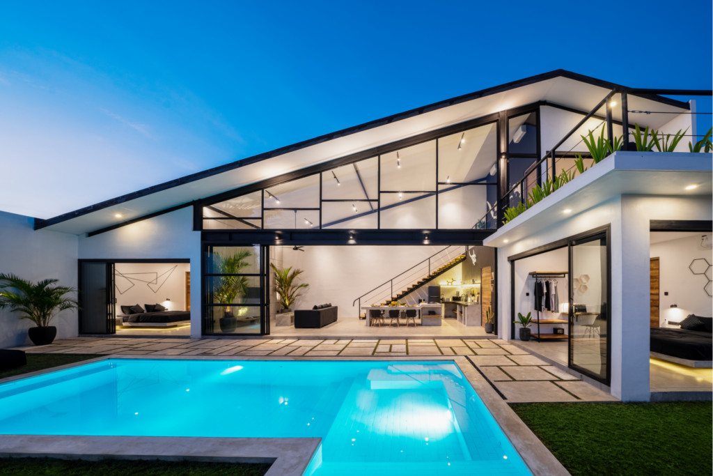 Modern, minimalist house with expansive glass walls and a bright, illuminated pool, showcasing open-concept interior design and outdoor living spaces.